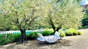 A garden with a bench under trees, a white picket fence, and a pool in the background.