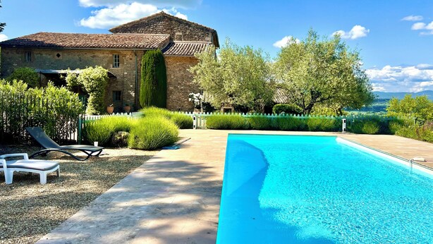 A stone building with a tiled roof, a swimming pool, and a well-maintained garden with a white picket fence.