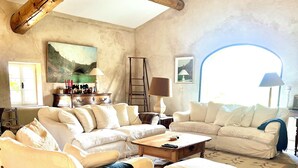 A spacious living room with a high ceiling and exposed wooden beams, featuring a large sectional sofa, a smaller sofa, and a coffee table.