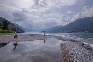 A pebbly beach with a calm lake, mountains in the background, and a cloudy sky.