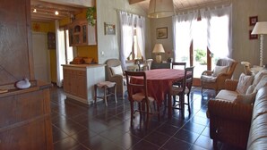 A spacious living area with a dining table, chairs, and a sofa. There is a kitchen area with cabinets and a window. - Villa Retreat with Pool and pets (Vélieux)
