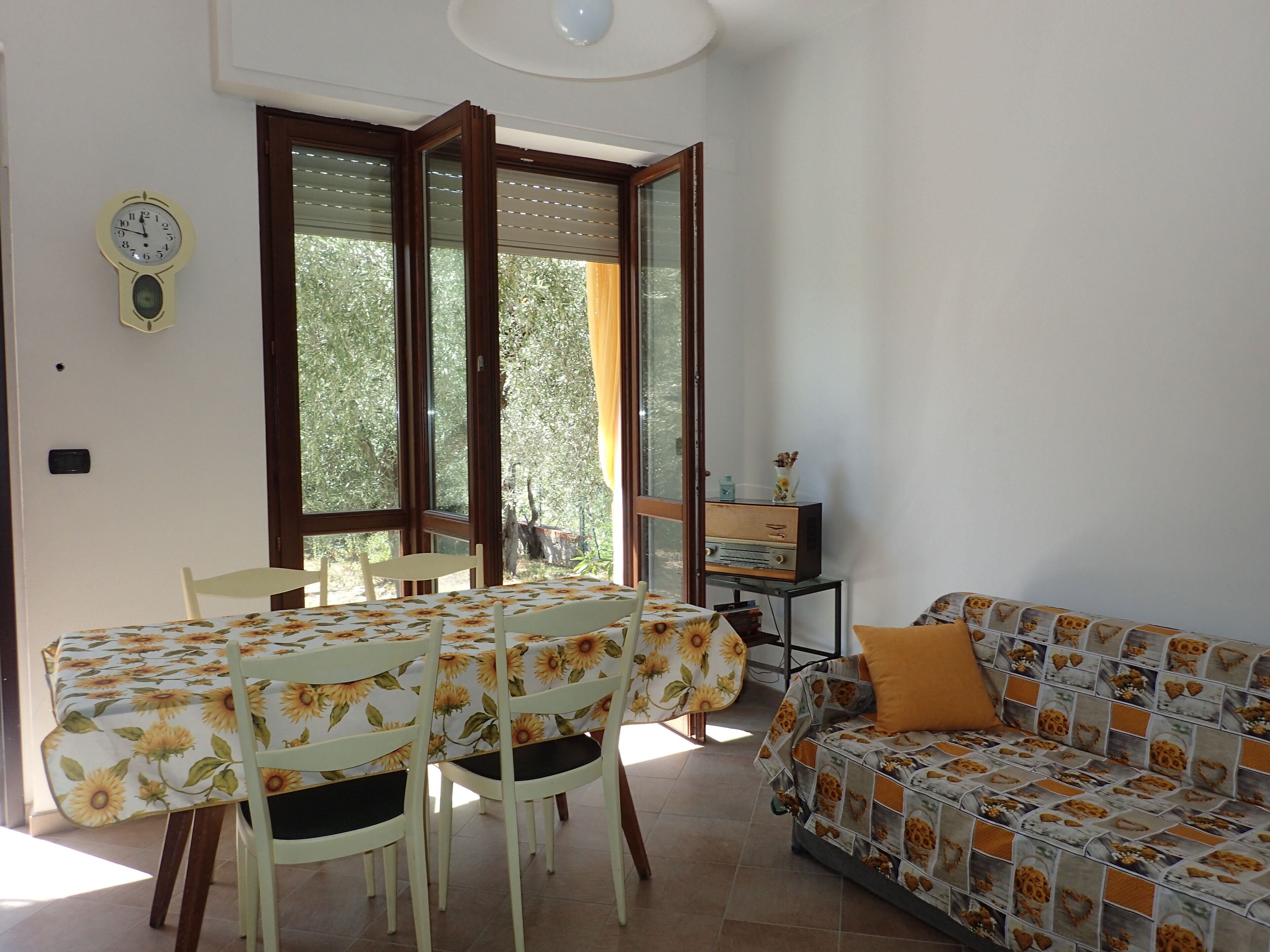 A dining area with a table and chairs, a sofa, a clock on the wall, and a window with blinds.