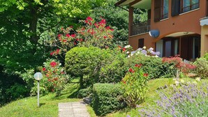 A two-story building with a balcony, surrounded by lush greenery and various flowering plants. - Italian Lake Apartment With Large Swimming Pool, Tennis Court, Lake Views, WiFi (Menaggio)