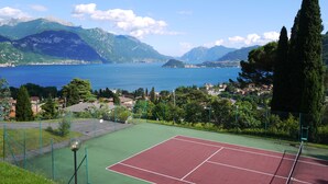 A tennis court with a net, surrounded by a fence and a green field. - Italian Lake Apartment With Large Swimming Pool, Tennis Court, Lake Views, WiFi (Menaggio)