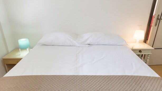 Ein Bett mit weißer Bettwäsche, zwei Kissen, einer beigen gesteppten Tagesdecke, einem Nachttisch mit einer blauen Lampe und einem weißen Beistelltisch mit einer Lampe.