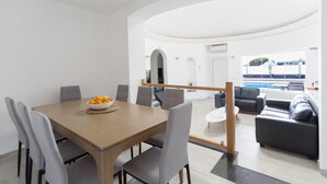 A modern dining area with a wooden table and grey chairs, a bowl of oranges on the table, and a view of a pool and outdoor seating area.