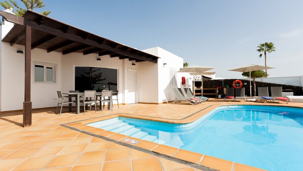 A swimming pool with a tiled deck, a covered patio area with chairs, and a white building with a door and windows.