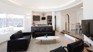 A modern living room with a black leather sofa, a white coffee table, and a black leather chair. There is a kitchen area in the background with a stone wall and a TV mounted above the counter.