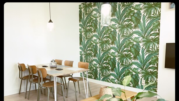 A dining area with a table and chairs, a hanging light fixture, and a wall with a tropical plant wallpaper.