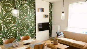 A living room with a sofa, a dining table with chairs, a TV, and a wall with a tropical leaf pattern.