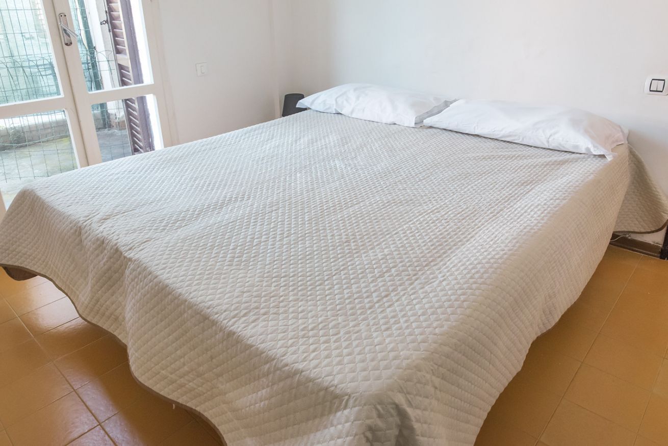 A bed with a quilted white cover, a window with blinds, and a tiled floor.