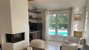 A living room with a fireplace, a television, a sofa, a coffee table, and a view of a swimming pool through a glass door. - A charming, 6-bedroom house in a convenient location (Gassin)