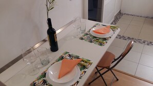 Ein Esstisch ist gedeckt mit Tellern, Gläsern und einer Flasche. Im Hintergrund steht ein Stuhl und eine Pflanze.