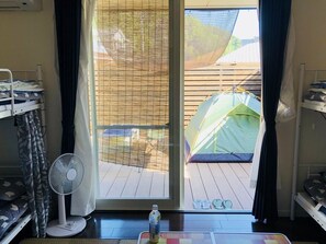 Una habitación con puerta de vidrio corredera que da a una terraza, un ventilador, una mesa con una botella y una tienda de campaña verde afuera.