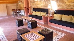 Un salon chaleureux avec un mur en pierre, un plafond en bois et un canapé agrémenté de coussins jaunes. On y trouve deux poufs noirs et une petite table en bois.