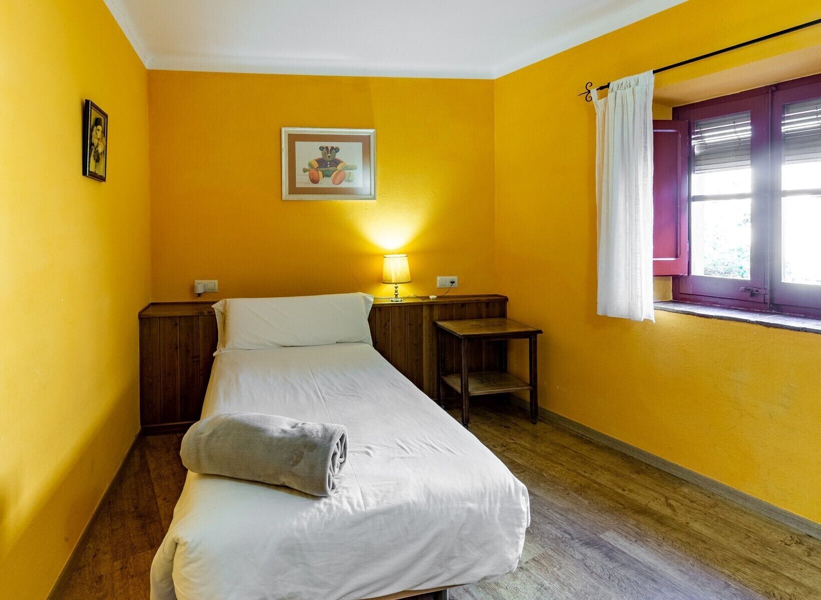 A small, single-bed room with a wooden headboard, a bedside table, a lamp, and a window with a curtain.