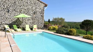 Un bâtiment en pierre doté d’une piscine, de fauteuils de jardin verts et offrant une vue sur la campagne.