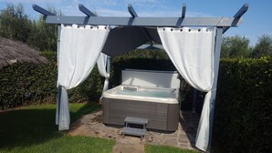 A hot tub under a pergola with white curtains, surrounded by greenery.