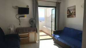 Una stanza con un divano blu, una televisione a parete, una ventola e vista mare attraverso una porta aperta.