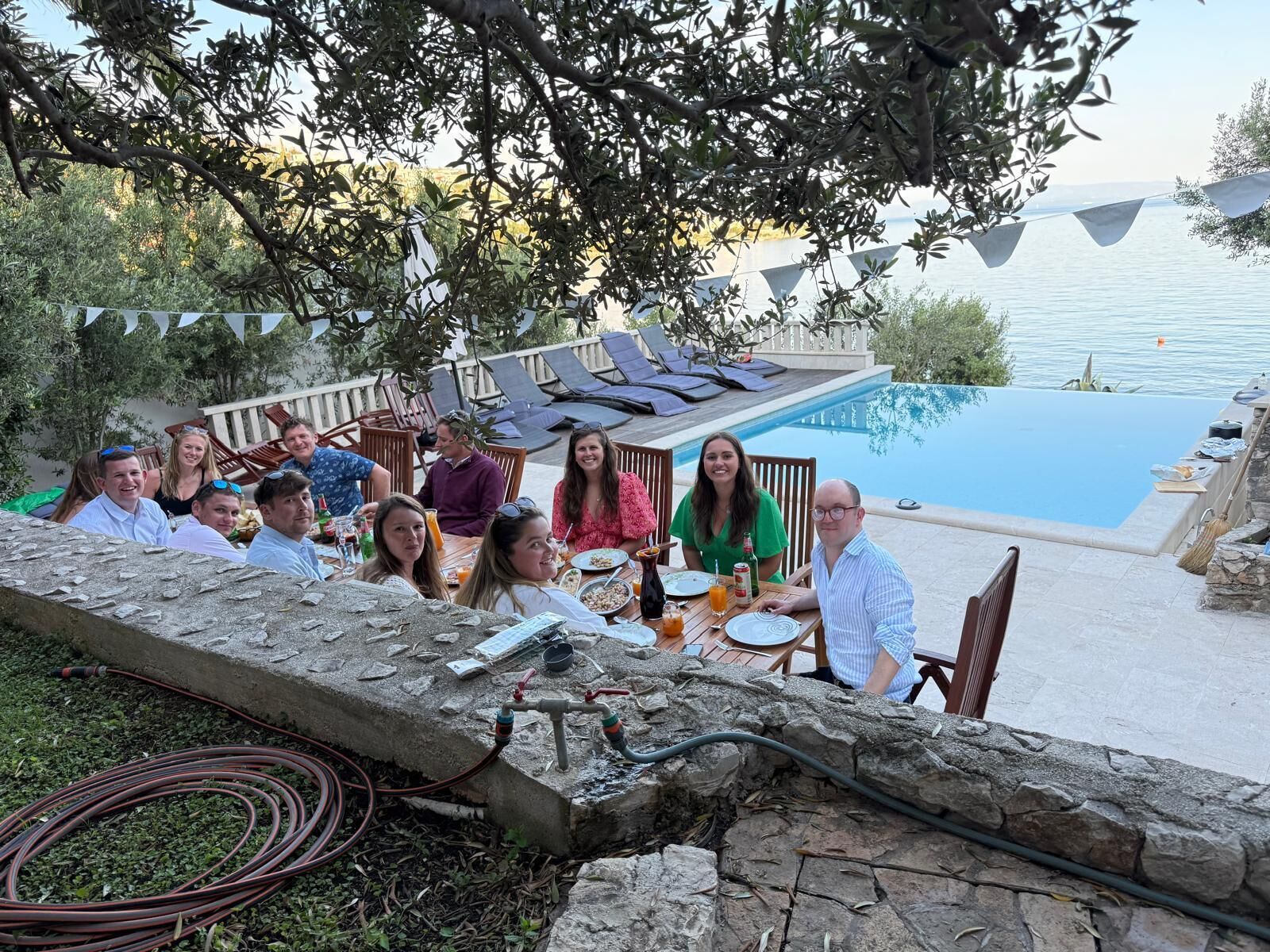 Un groupe de personnes est assis à une table en plein air, près d’une piscine, et profite de la vue sur l’eau et d’une terrasse aménagée avec des chaises longues.