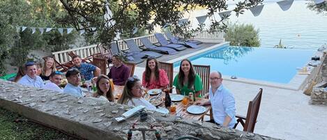 Un groupe de personnes est assis à une table en plein air, près d’une piscine, et profite de la vue sur l’eau et d’une terrasse aménagée avec des chaises longues.