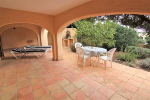 Eine überdachte Terrasse mit Tisch, Stühlen und einer Liege, mit Blick auf den Garten.
