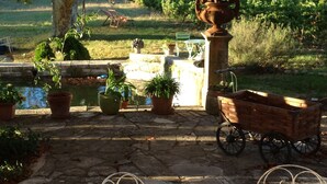 Un patio de piedra con una mesa y sillas, rodeado de plantas en macetas y un carro rústico.