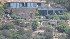 Une maison à la façade en pierre, dotée de grandes fenêtres et d’un toit en tuiles, entourée de verdure et de rochers.