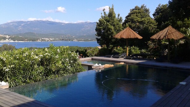 Une piscine avec vue sur la mer et les montagnes, entourée de verdure et agrémentée de deux parasols en chaume.