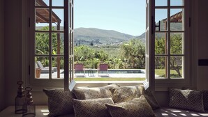 A room with a large window overlooking a pool and mountains. There are several patterned cushions on a bench. - Classic Three Bedroom Cottage with private pool (Zakynthos)