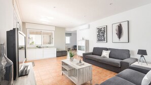 A modern living room with a grey sofa, a coffee table, and a TV. There is a kitchen area with white cabinets and a dining table. - Villa La Calma - beach,private Pool, free AC&WiFi (Es Mercadal)