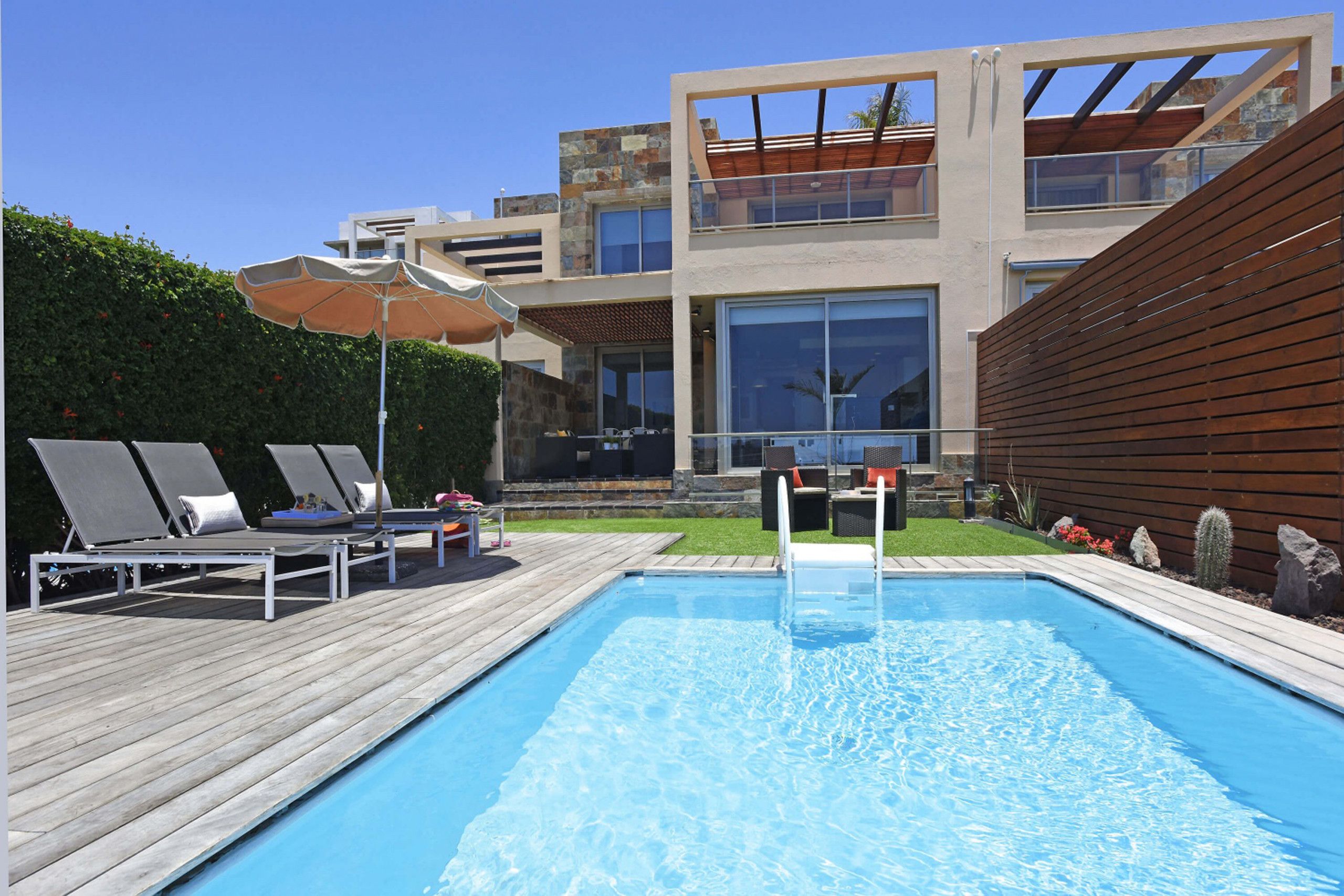 Un área de piscina moderna con sillones de descanso y sombrillas, rodeada por una terraza de madera y un jardín bien cuidado.