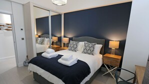 A modern hotel room with a large bed, two bedside tables with lamps, a mirror, and a chair. - Apartment 2 Waterstone House - Sea View Apartment (Tenby)