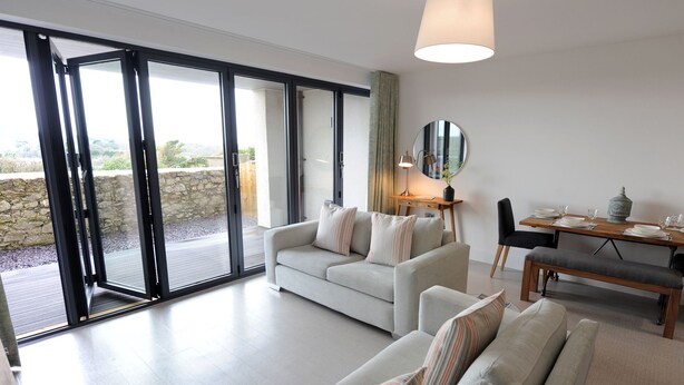 A modern living room with a sofa set, a dining table with chairs, and a large sliding glass door leading to an outdoor area. - Apartment 2 Waterstone House - Sea View Apartment (Tenby)