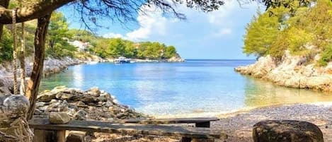 A bench by a clear, calm body of water with rocky shores and trees in the background.