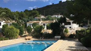 A swimming pool with a stone border, surrounded by a garden with various plants and a hillside with houses in the background. - Detached 2 Bedroom Villa in El Portet, with large terraces and pool views (Teulada)