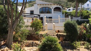 A white villa with a balcony, surrounded by a garden with palm trees and potted plants. - Detached 2 Bedroom Villa in El Portet, with large terraces and pool views (Teulada)