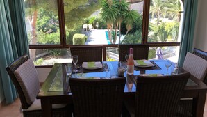 A dining area with a table set for four, wicker chairs, and a view of a garden through a large window. - Detached 2 Bedroom Villa in El Portet, with large terraces and pool views (Teulada)