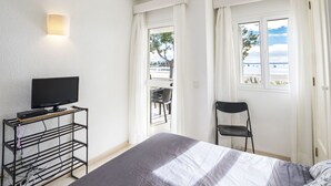 A hotel room with a bed, a TV on a stand, a chair, and a window with a view of the sea. - Cosy apartment for 4 people with WIFI, A/C, TV and patio (Alcúdia)