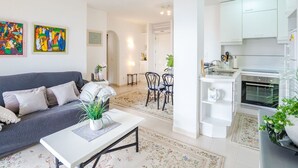 A modern living room with a grey sofa, a white coffee table, and a dining area with a table and chairs. A kitchen with white cabinets and appliances is visible in the background. - Cosy apartment for 4 people with WIFI, A/C, TV and patio (Alcúdia)