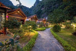 A pathway leading to traditional-style buildings surrounded by lush greenery and a mountainous backdrop.