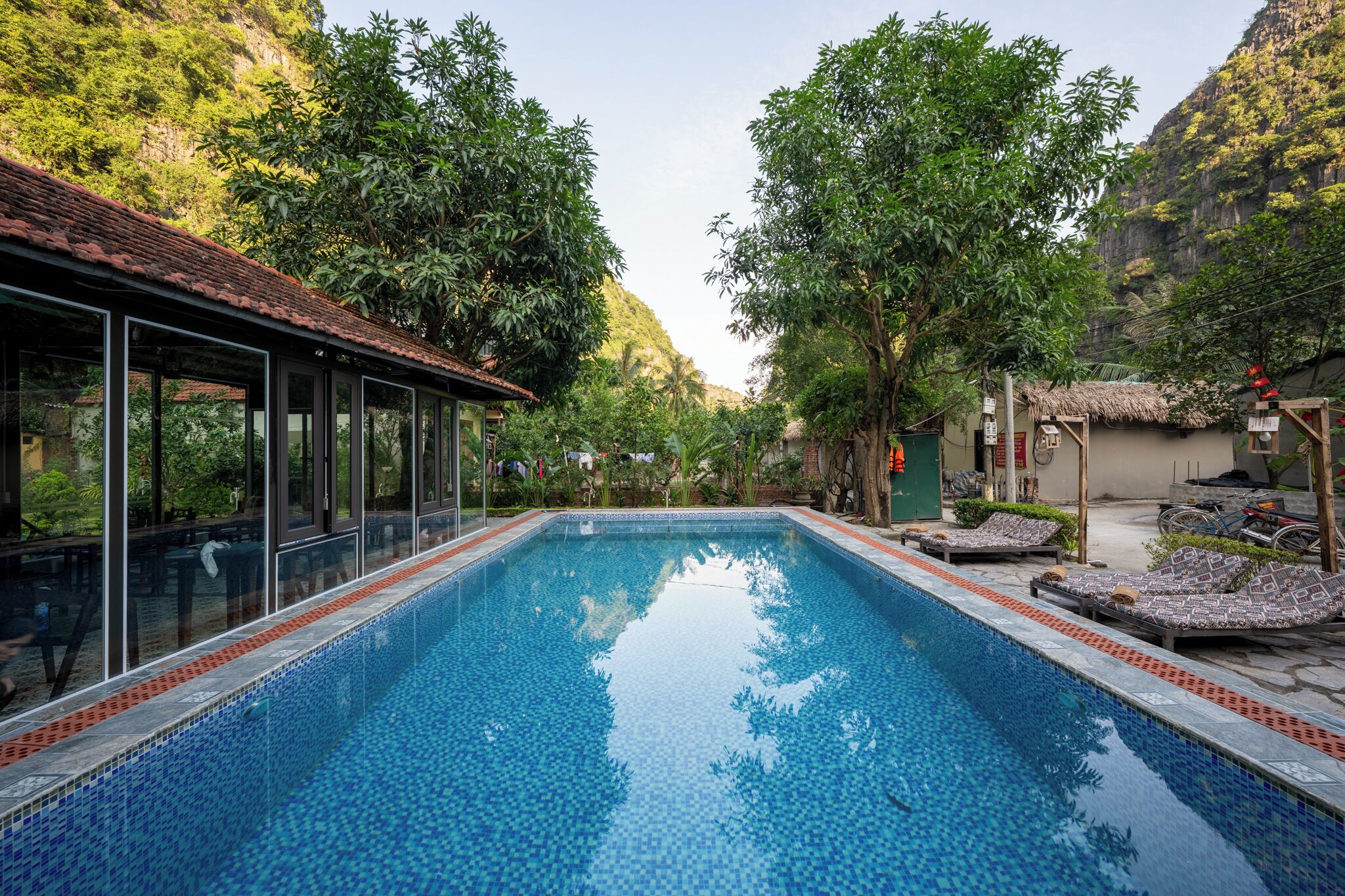 Una piscina rectangular de agua cristalina, rodeada de abundante vegetación y con montañas al fondo.