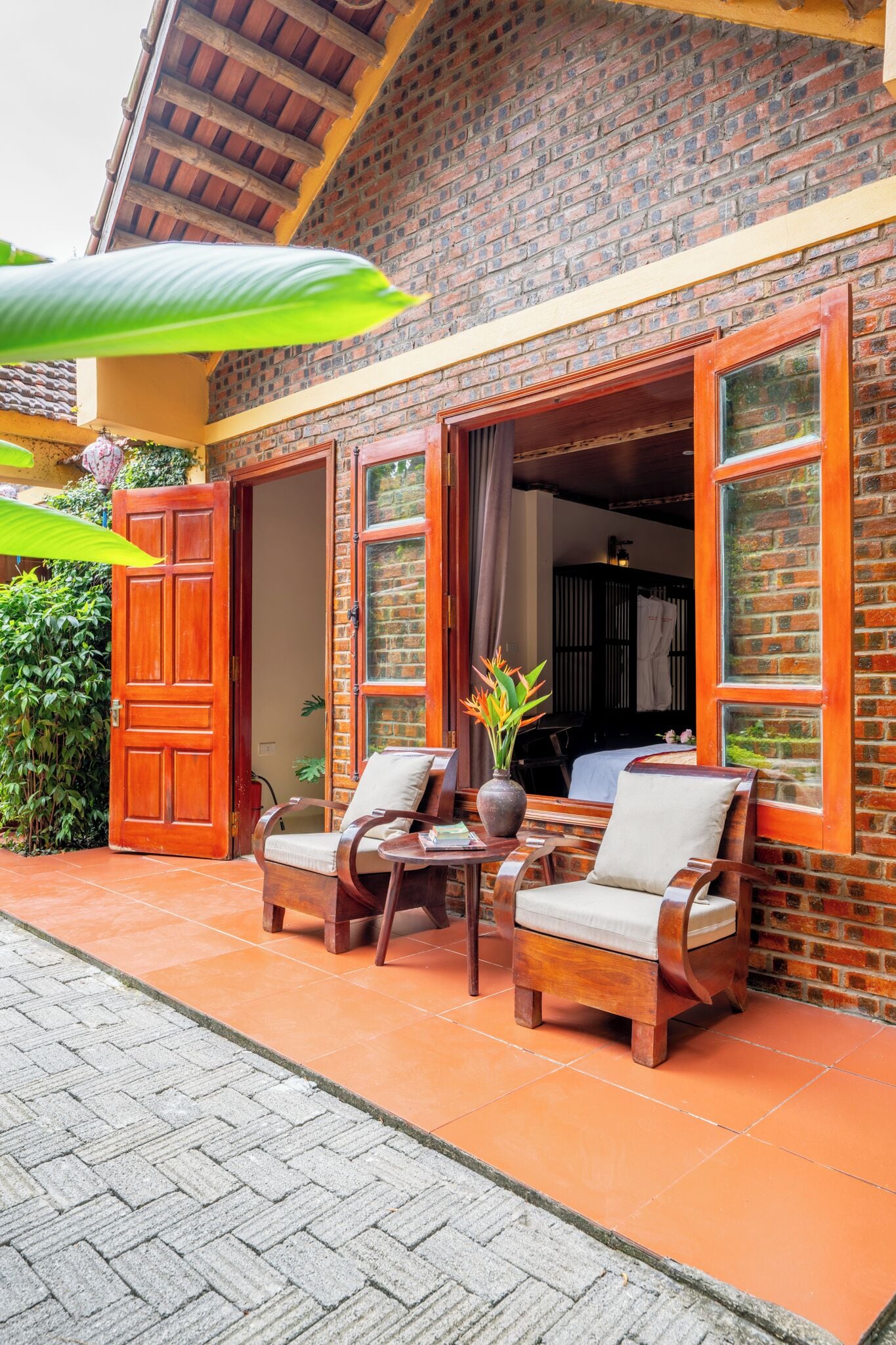 A brick building with open doors leading to a room, two wooden chairs with cushions, a potted plant, and a tiled patio area.