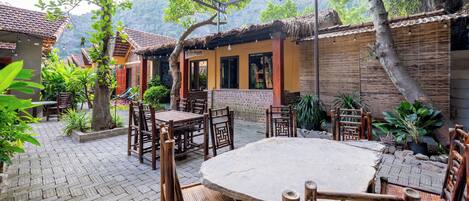Un espacio exterior rústico para comer con mesas y sillas de madera, rodeado de plantas en macetas y una valla de bambú.