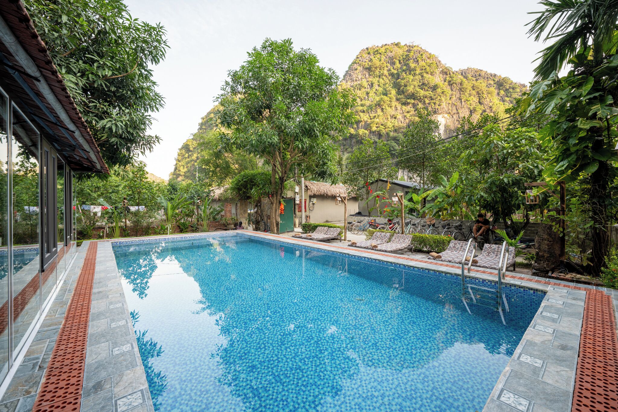 A rectangular swimming pool with a tiled edge, surrounded by lush greenery and a mountain backdrop.