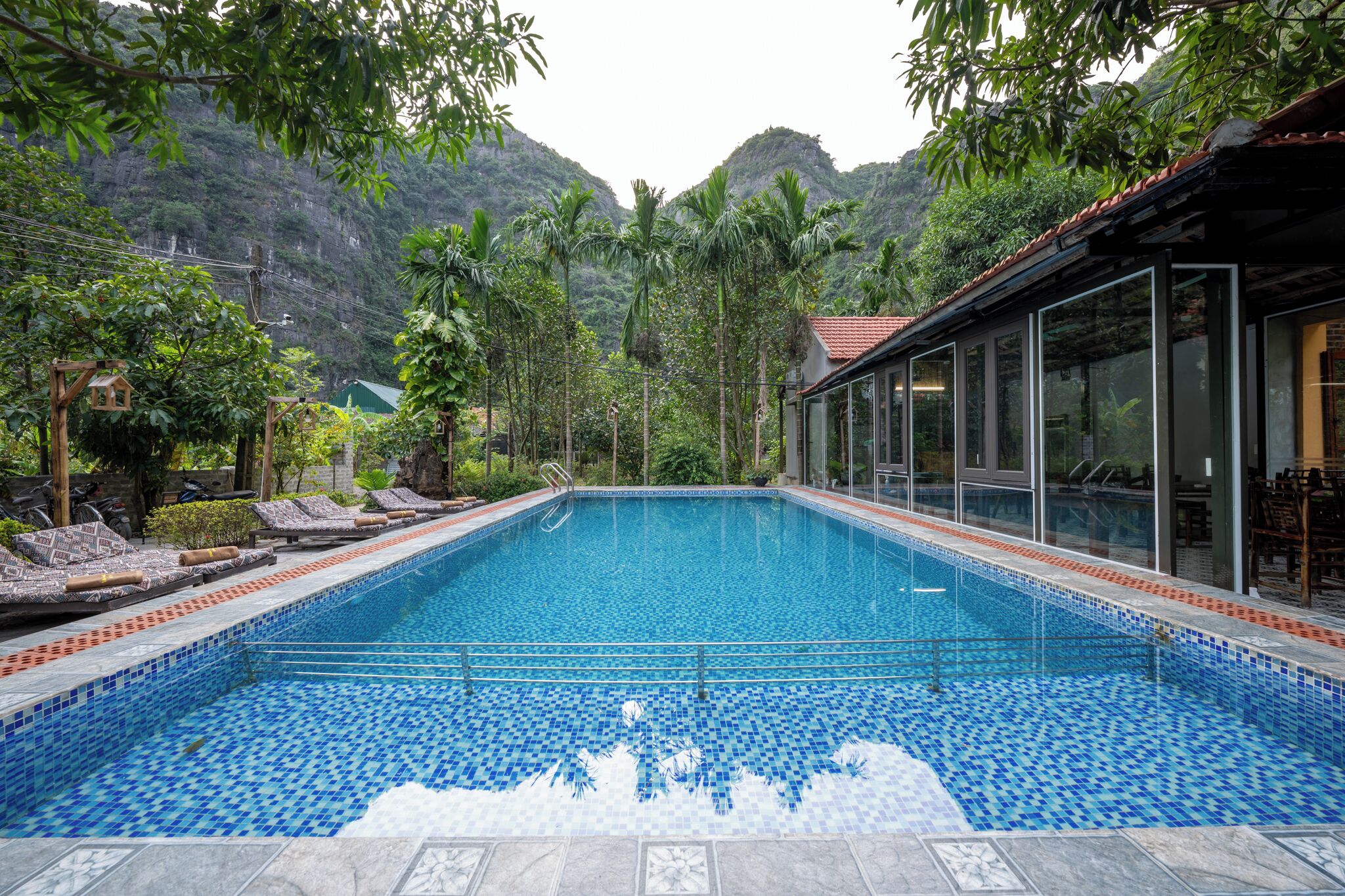 A rectangular swimming pool with blue mosaic tiles, surrounded by lounge chairs and a glass-enclosed area with tables and chairs.