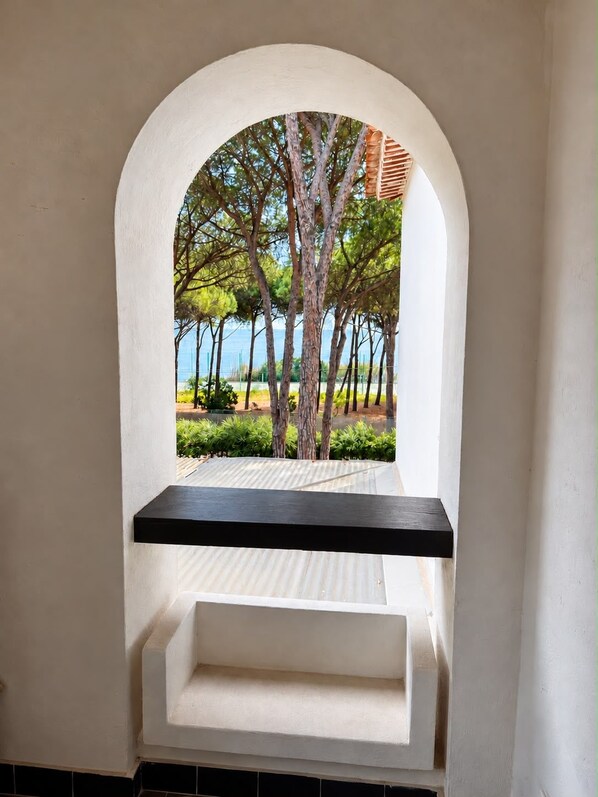 A white arched window with a black shelf, overlooking a landscaped area with trees and a view of the sea.