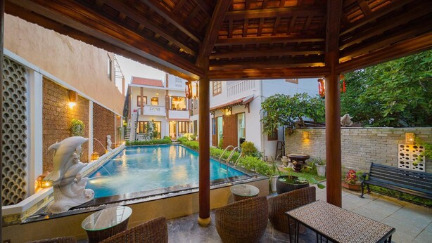 A covered outdoor pool area with wicker furniture, a statue, and a building in the background. - Green Grapefruit Villa (Da Nang)