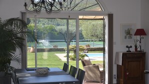 Comedor con una mesa y sillas, una planta en maceta y vistas a una cancha de tenis y una piscina a través de un ventanal.