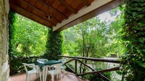 Une terrasse couverte, agrémentée d’une table et de chaises, entourée d’une végétation luxuriante et d’une rambarde en bois.